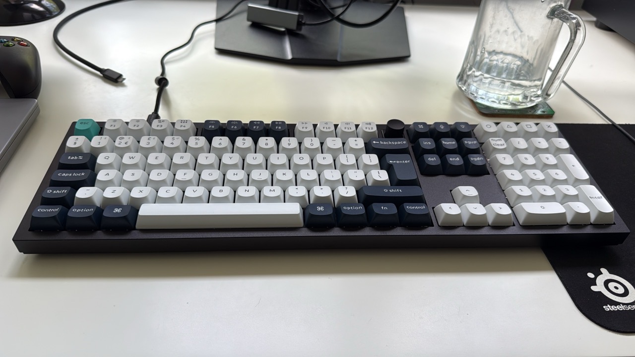 A black and white keyboard sitting on a white desk with a black mouse mat and a glass of water