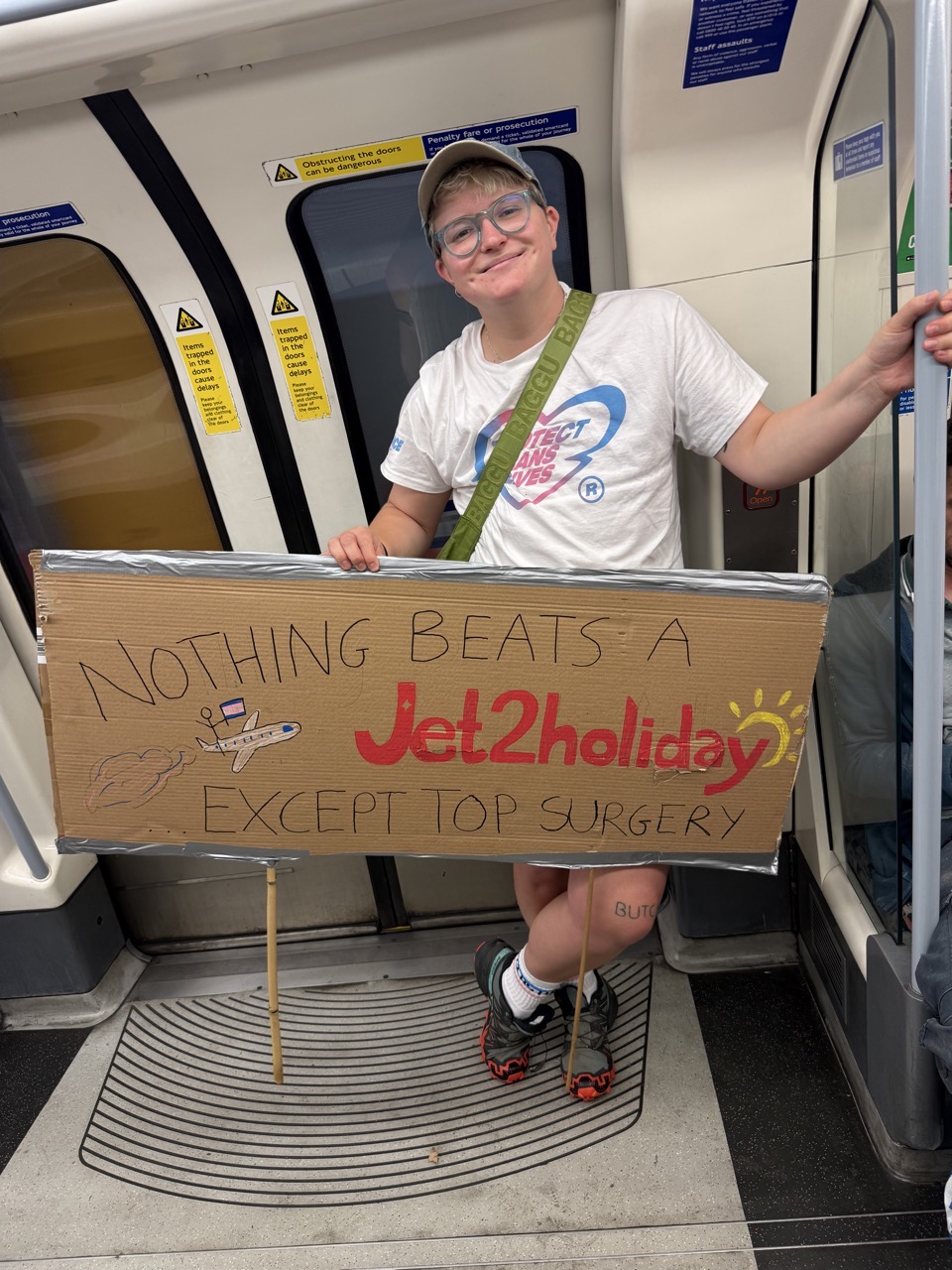 My very cool friend Vincent on the tube with a sign that says Nothing beats a Jet2 holiday except top surgery