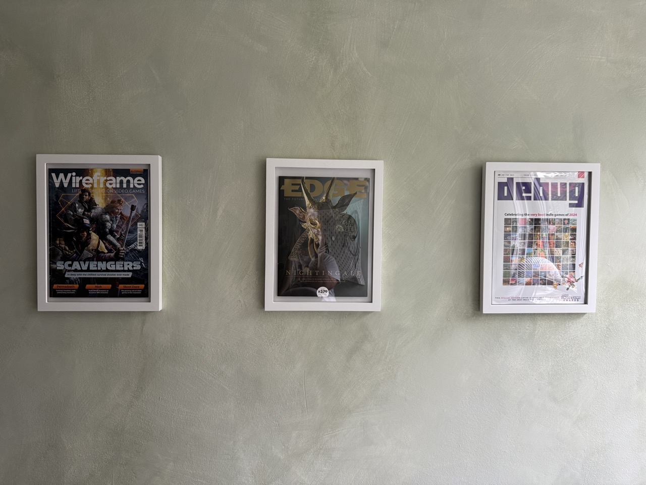Three frames on a wall with magazines. Scavengers on Wireframe, Nightingale on Edge, and the Debug Awards cover with Feud